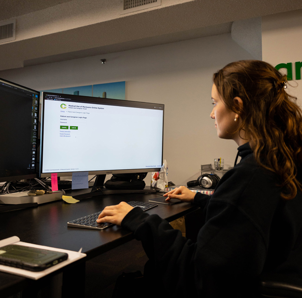 The image shows a jd software employee sitting at a desk, working on a computer with a large monitor displaying the Medical Use of Marijuana Online System.